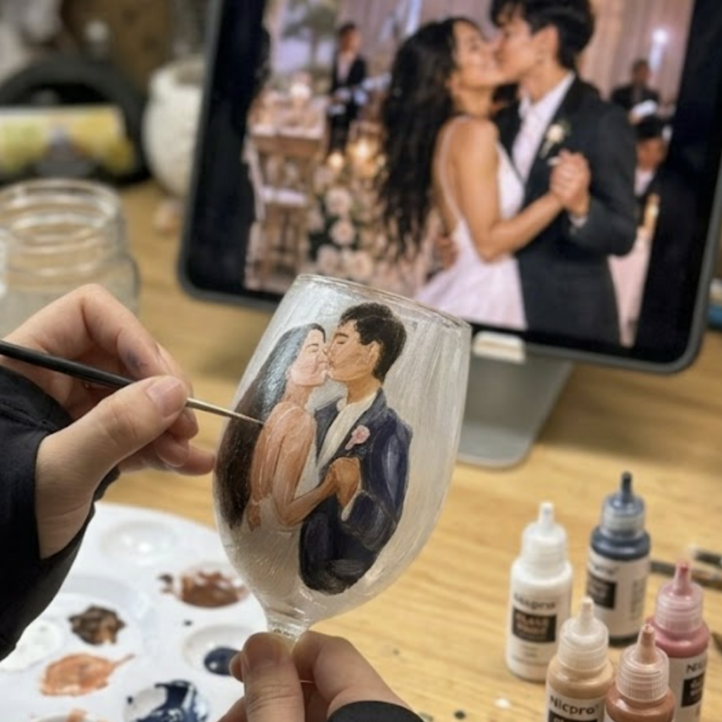 Artist hand-painting a bride and groom's wedding portrait onto a wine glass, using a photograph displayed on a tablet as reference.
